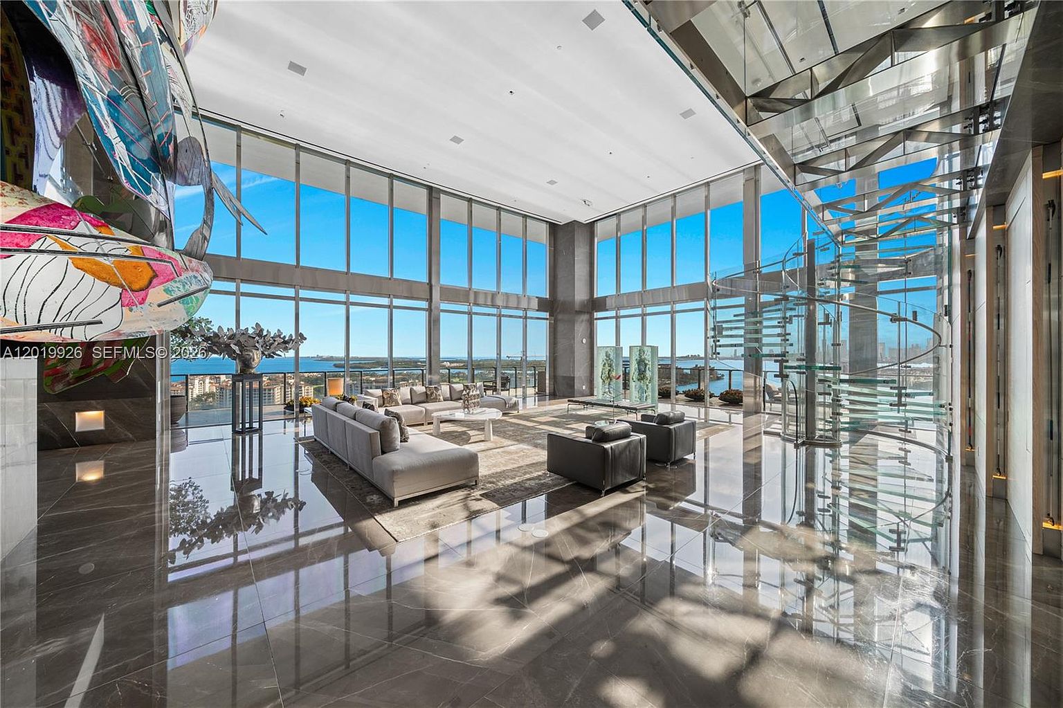 This expansive, ultra-modern living room features polished stone flooring and dramatic floor-to-ceiling windows that offer panoramic water views. The space is anchored by a large, neutral-toned sectional sofa and a glass-railed spiral staircase, creating a sophisticated and airy atmosphere. A vibrant, abstract sculpture stands prominently in the foreground, adding a bold artistic touch to the minimalist, high-ceilinged interior.