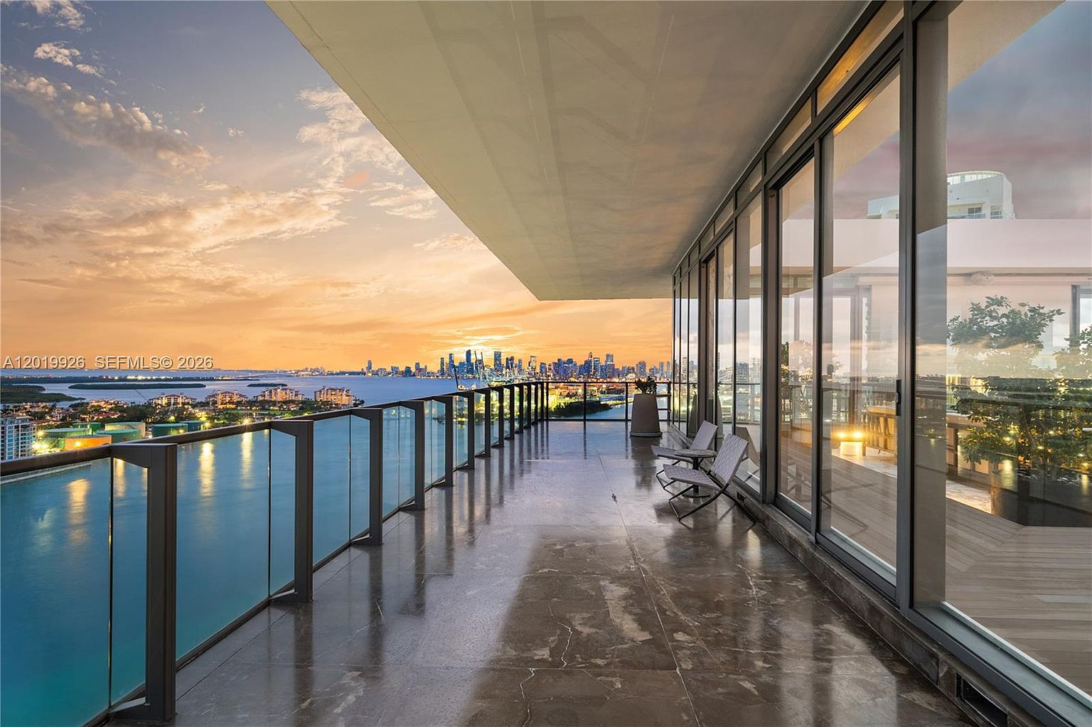 This expansive, covered balcony offers a panoramic view of a city skyline across a body of water at sunset. The space features polished stone flooring, a sleek glass railing, and floor-to-ceiling glass walls that provide seamless indoor-outdoor connectivity. Two modern chairs are positioned to take advantage of the dramatic horizon, creating a sophisticated and serene atmosphere.