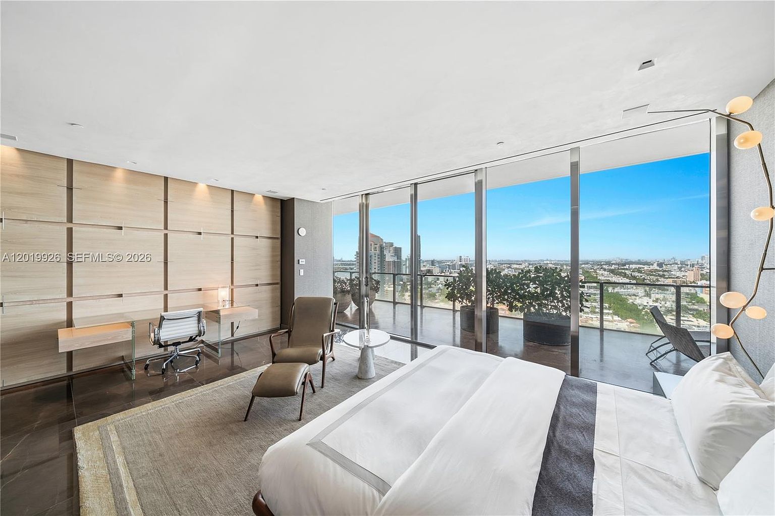 This spacious primary bedroom features a minimalist aesthetic with a large bed centered in the room and a dedicated workspace against a light wood-paneled wall. Floor-to-ceiling windows provide an expansive, unobstructed view of the city skyline and surrounding landscape, flooding the room with natural light. The space is accented by a modern, sculptural floor lamp and a neutral-toned area rug, creating a sophisticated and serene atmosphere.