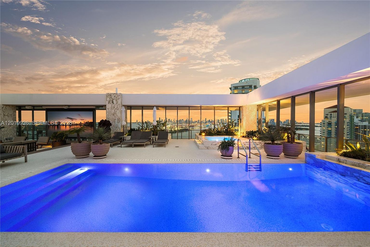 This luxurious rooftop terrace features a vibrant blue swimming pool in the foreground and a separate spa area set against a backdrop of a sprawling city skyline. The space is furnished with lounge chairs and potted plants, framed by expansive glass walls that offer panoramic views during a golden sunset. The modern design emphasizes an open-air living experience, blending high-end amenities with a dramatic urban perspective.