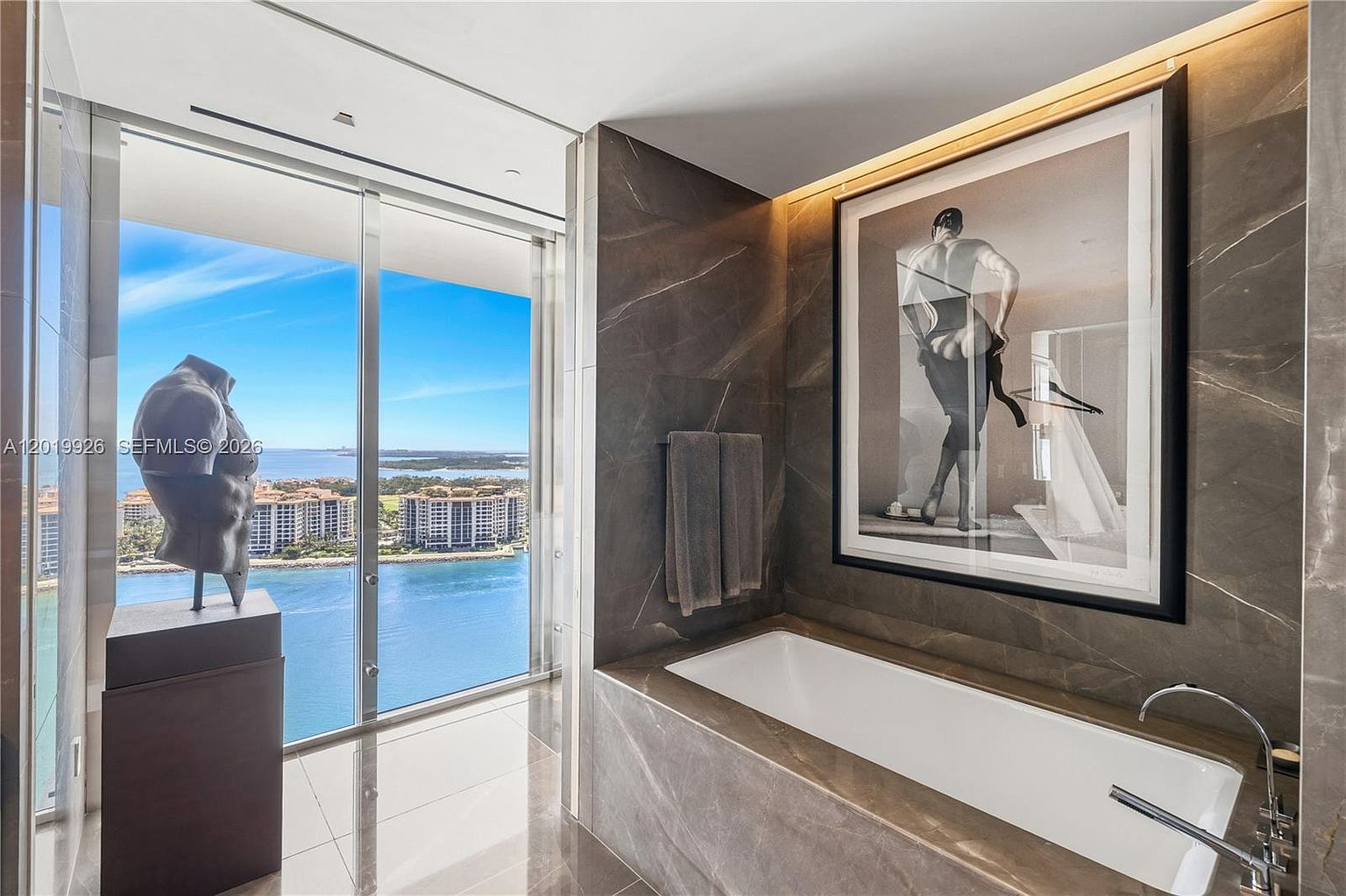 This luxurious primary bathroom features a deep soaking tub set against a backdrop of dark, veined stone walls. A large, framed art piece hangs above the tub, while floor-to-ceiling windows provide an expansive, panoramic view of the water and surrounding landscape. A sculptural bust on a pedestal stands near the glass, enhancing the sophisticated and artistic atmosphere of the space.