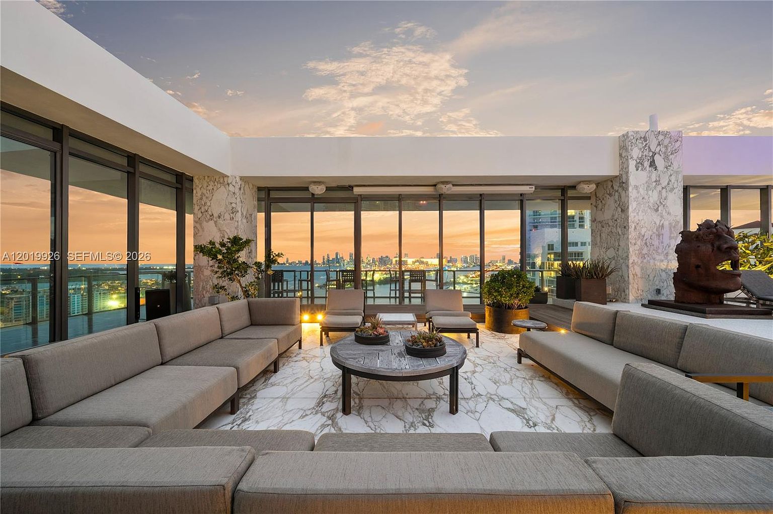 This expansive, high-end terrace offers a sophisticated outdoor living space with a panoramic view of the city skyline at sunset. The area is furnished with a large, U-shaped sectional sofa and a central circular coffee table, all set upon a patterned stone floor. Large glass panels provide a seamless transition to the outdoors, while the open-air design creates an airy, luxurious atmosphere perfect for entertaining.