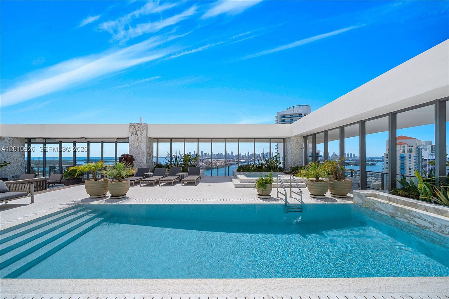 This high-rise rooftop terrace features a pristine swimming pool as its centerpiece, set against a backdrop of expansive city skyline and water views. The deck is furnished with multiple lounge chairs and potted plants, framed by large glass panels that provide an unobstructed panoramic vista. The scene conveys a luxurious, modern lifestyle with a bright, open-air atmosphere.