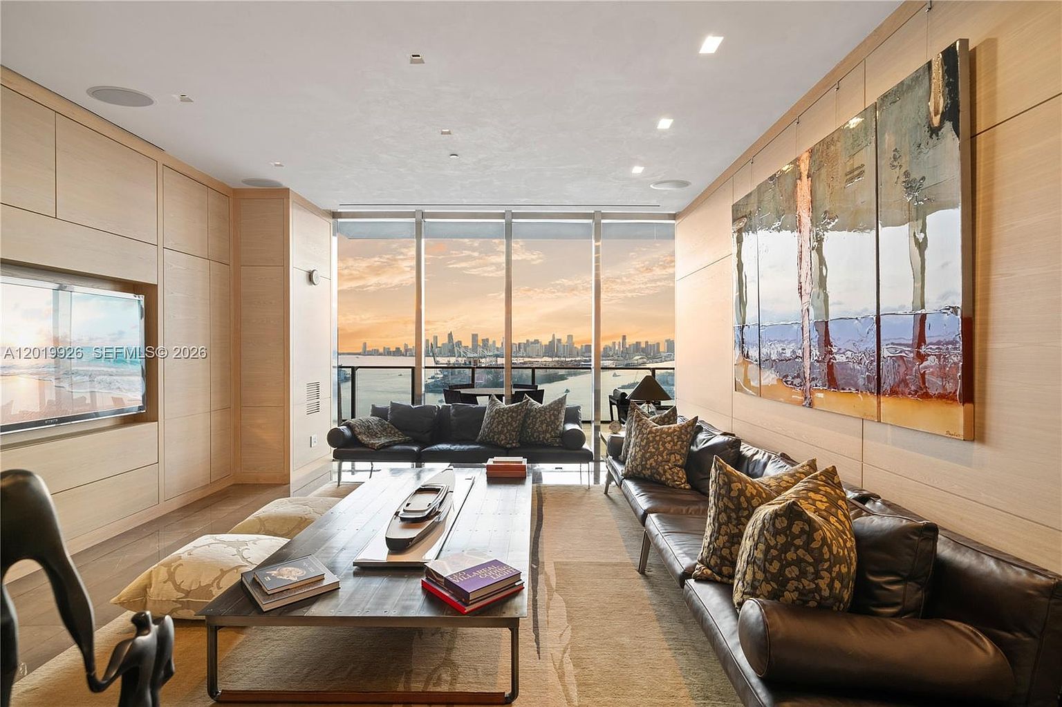This sophisticated living room features light wood wall paneling and a large, dark leather sofa arrangement centered around a long, low-profile wooden coffee table. The space is anchored by floor-to-ceiling windows that offer a panoramic view of a city skyline and waterfront at sunset. A large, multi-panel abstract painting hangs on the right wall, adding a vibrant artistic focal point to the contemporary interior.