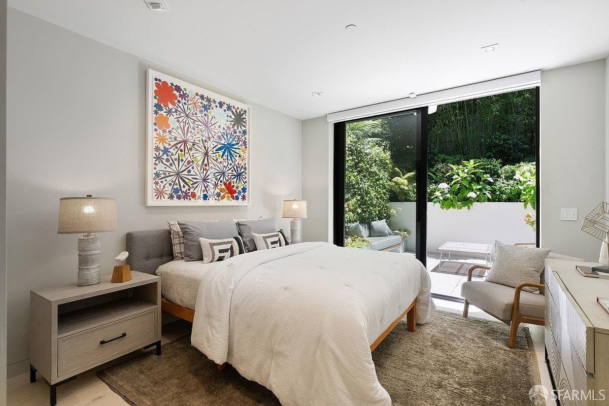 This contemporary bedroom features a neutral color palette, a platform bed with a grey upholstered headboard, and a vibrant, multi-colored abstract art piece above the bed. Large sliding glass doors provide seamless access to a private outdoor patio, allowing for abundant natural light and a view of lush greenery. The room is furnished with light wood nightstands and a comfortable armchair, creating a serene and inviting atmosphere.