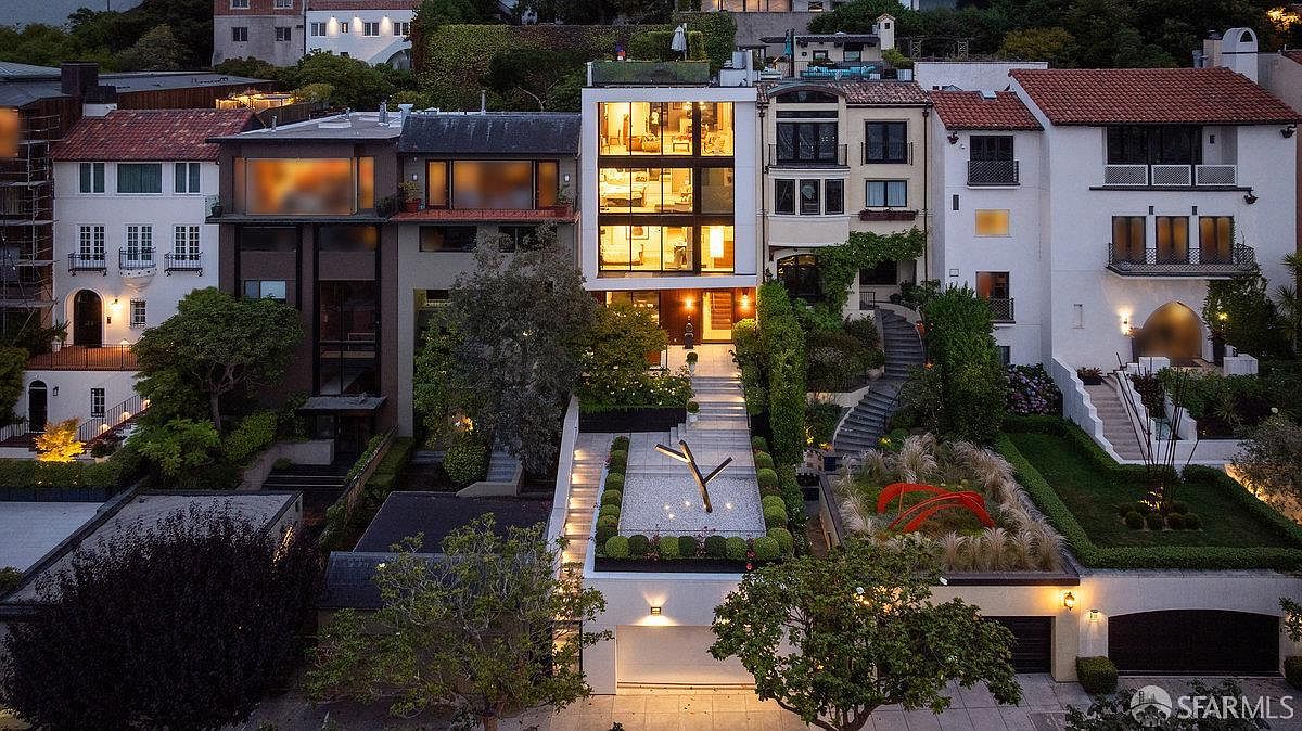 This aerial perspective captures a row of upscale, multi-story residences nestled on a hillside, showcasing a blend of modern and traditional architectural styles. The central home features a striking glass-fronted facade with illuminated levels, leading down to a tiered landscape with a sculptural art piece and manicured greenery. The composition highlights the sophisticated urban design and the terraced outdoor spaces that define this luxury property.