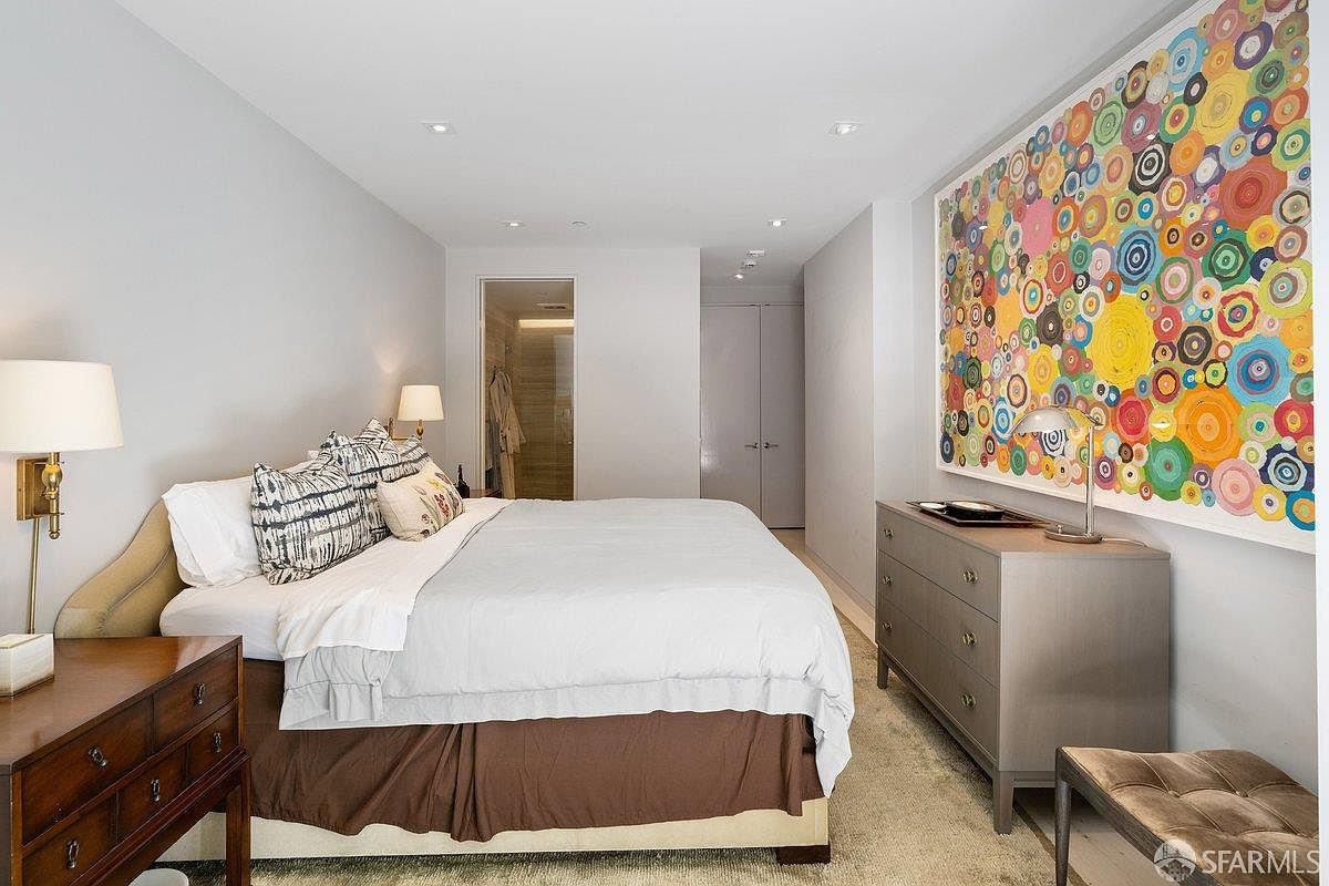 This primary bedroom features a neutral color palette accented by a large, vibrant abstract art piece on the wall. The room is furnished with a plush bed topped with patterned pillows, a dark wood side table, and a sleek gray dresser. A doorway in the background leads to an en-suite bathroom, creating a cohesive and well-lit living space.