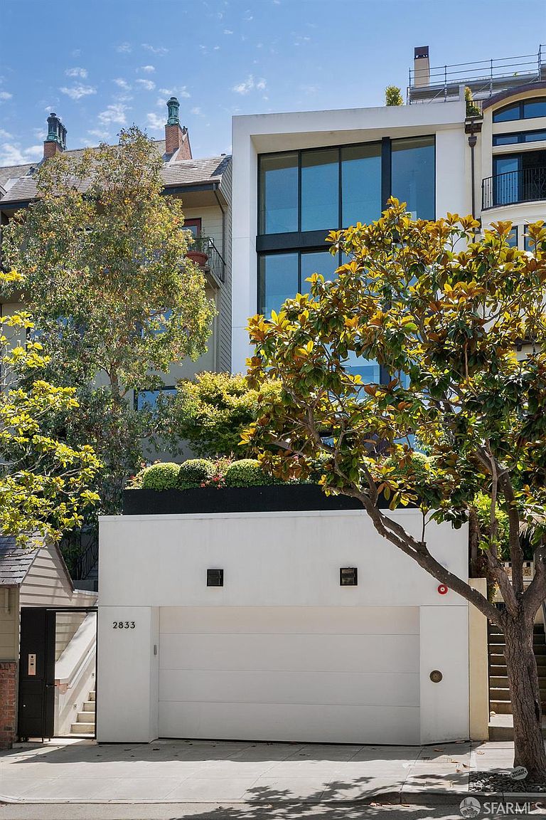 This modern residential facade features a prominent white garage structure at street level, topped by a landscaped terrace with lush greenery. Above the garage, a multi-story section showcases large windows that span the width of the building, offering a contemporary contrast to the more traditional neighboring homes. The perspective is a straight-on street view, highlighting the clean lines and architectural integration of the property within its urban setting.