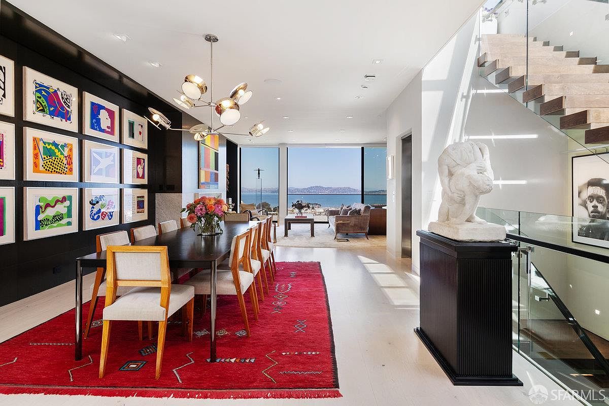 This sophisticated dining area features a dark wood table set against a dramatic black accent wall adorned with a gallery of colorful framed artwork. A vibrant red patterned rug anchors the space, while a modern multi-bulb chandelier hangs above the table, leading the eye toward the expansive glass wall that frames a panoramic water view. To the right, a sculptural piece rests on a dark pedestal near a floating staircase with glass railings, creating a seamless blend of art and architecture.