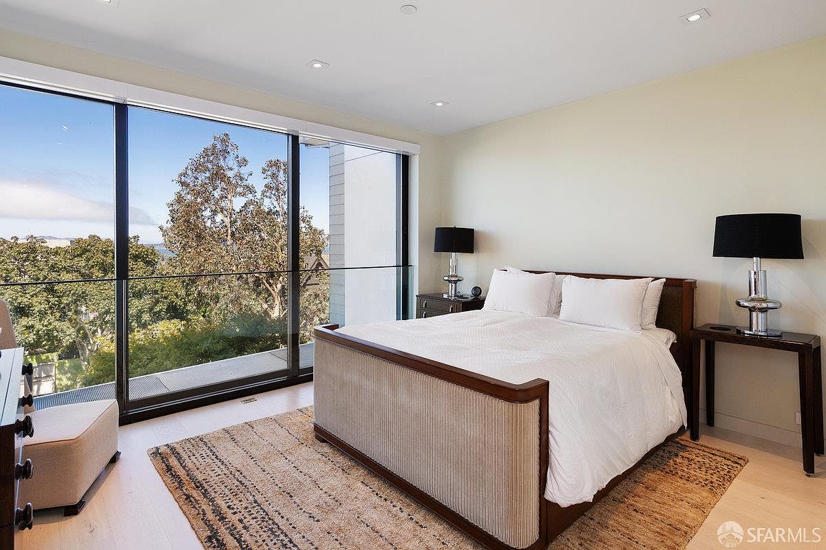 This bright and airy primary bedroom features a large bed with a textured headboard and crisp white bedding, centered in the room. A wall of floor-to-ceiling windows provides an expansive view of the lush green trees outside, filling the space with natural light. The room is accented by two matching bedside tables with modern lamps and a woven area rug, creating a serene and contemporary atmosphere.