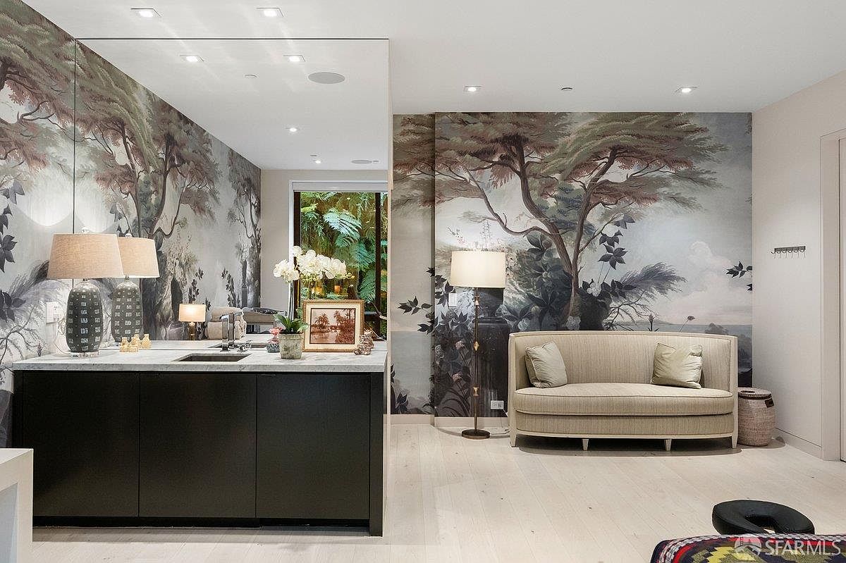 This sophisticated primary bathroom features a striking mural wallpaper depicting a serene forest scene that extends across the wall behind a neutral-toned sofa. A dark, sleek vanity with a stone countertop and integrated sink sits to the left, complemented by a large mirror that reflects the lush greenery visible through the window. The space is illuminated by warm floor and table lamps, creating a tranquil and luxurious atmosphere with light-toned flooring throughout.