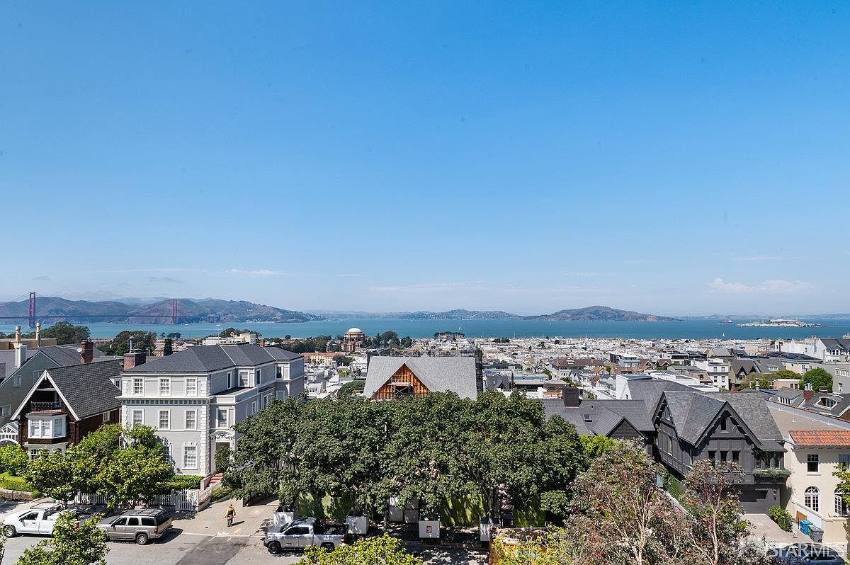 This elevated perspective captures a sweeping panoramic view of a dense urban neighborhood, showcasing a mix of historic and contemporary residential architecture. In the distance, the iconic Golden Gate Bridge spans the bay under a clear blue sky, providing a dramatic backdrop to the cityscape. The foreground features lush green trees and a quiet street, emphasizing the property's prime location with expansive vistas.