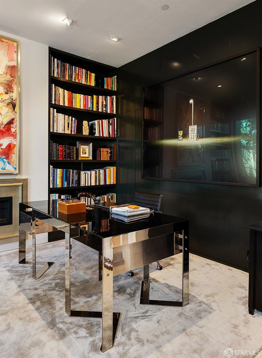 This sophisticated home office features a sleek, modern desk with a polished metal frame and a black glass top, positioned against a wall of dark, built-in shelving filled with books. To the left, a vibrant, multi-colored abstract painting adds a bold pop of color, while a large, dark-framed mirror on the right wall reflects the room's interior. The space is grounded by a soft, textured area rug, creating a professional yet refined atmosphere.