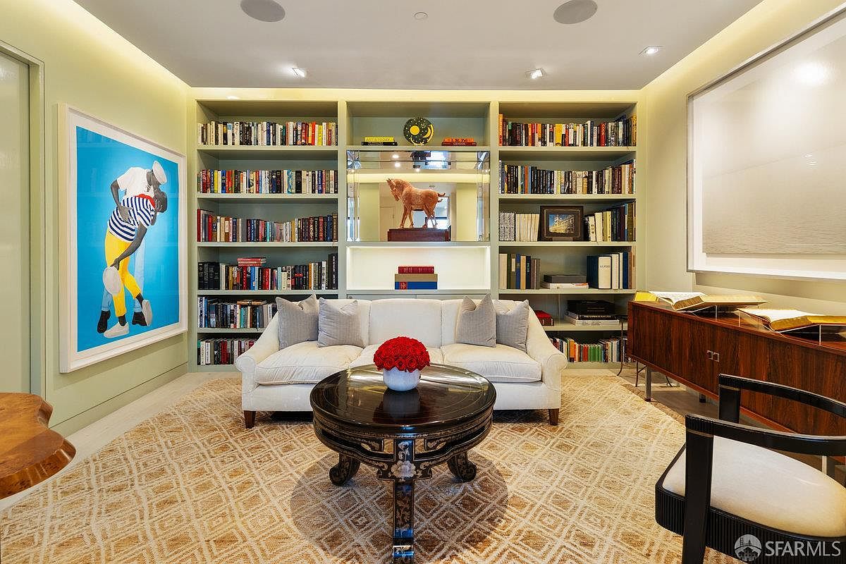 This sophisticated study or library features built-in shelving flanking a central mirrored display area, creating a symmetrical and elegant focal point. A plush white sofa sits before a dark, ornate round coffee table, while a vibrant piece of modern art adds a bold splash of color to the light-toned walls. The room is grounded by a textured, patterned area rug and accented by a dark wood console table, offering a refined and scholarly atmosphere.