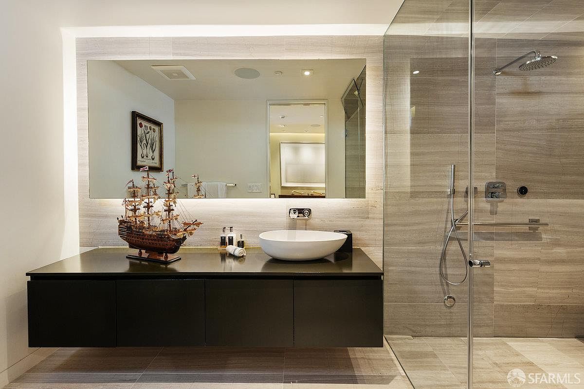 This modern primary bathroom features a sleek, floating dark vanity topped with a white vessel sink and a decorative model ship. A large, frameless mirror spans the wall above the vanity, complemented by warm under-cabinet lighting and a walk-in shower with glass paneling and stone-tiled walls. The perspective is a straight-on shot that highlights the clean lines and contemporary aesthetic of the space.