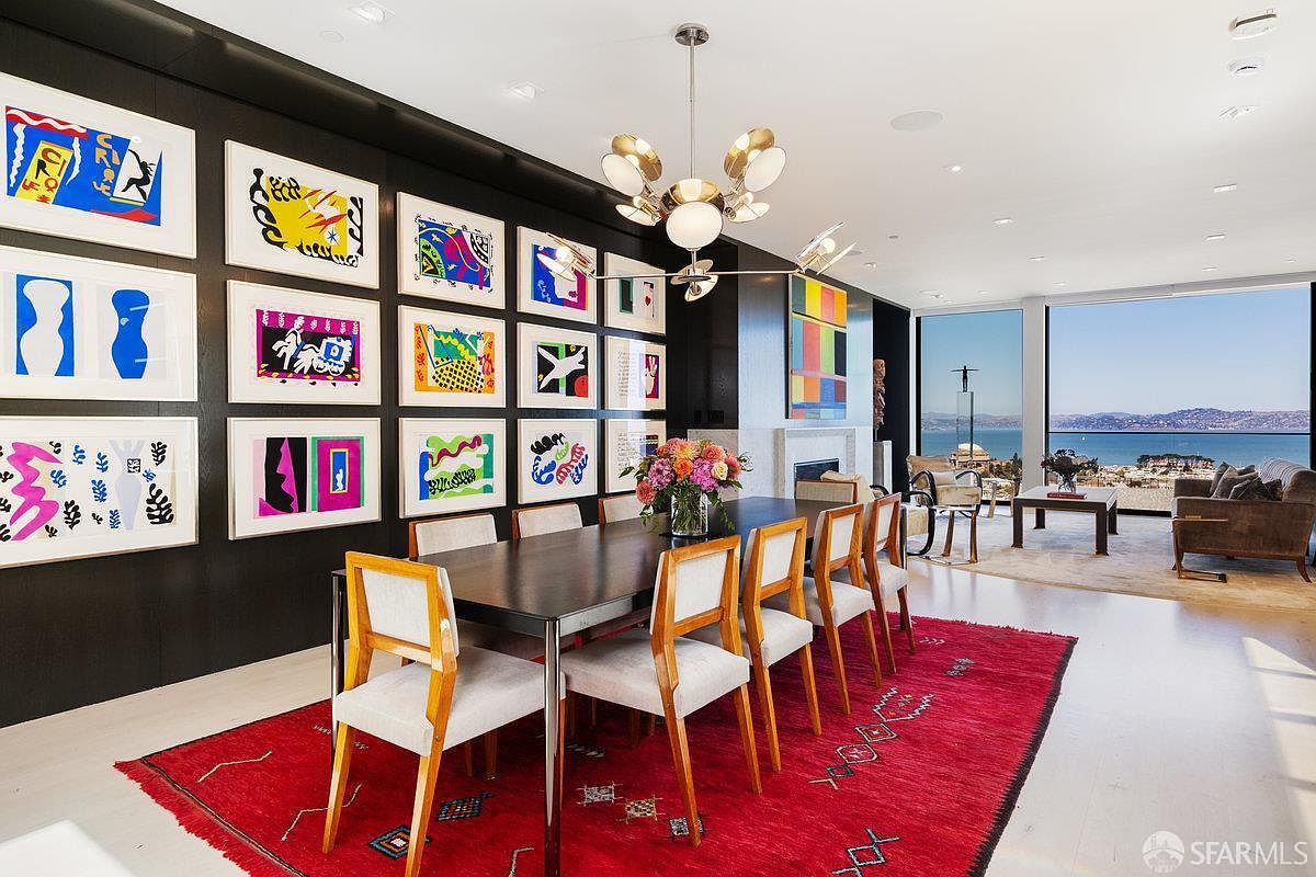 This sophisticated dining room features a dark, gallery-style wall adorned with a grid of colorful abstract art prints, contrasting with a bright, open-concept living area. A long dining table with light-toned chairs sits atop a vibrant red patterned rug, while a modern, multi-globe chandelier hangs above. Large windows provide an expansive, panoramic view of the bay and distant hills, creating a seamless connection between the refined interior and the scenic outdoor landscape.