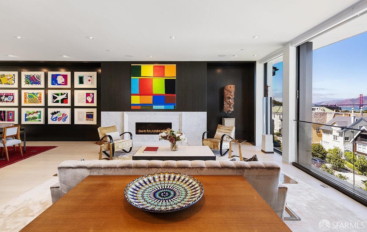This sophisticated living room features a striking gallery wall of colorful artwork set against a dark accent wall, centered around a fireplace with a white stone surround. The space is furnished with two modern armchairs and a low-profile coffee table, while a large, decorative bowl sits on a foreground table. Expansive floor-to-ceiling windows provide a panoramic view of the Golden Gate Bridge and the surrounding neighborhood, bathing the room in natural light.