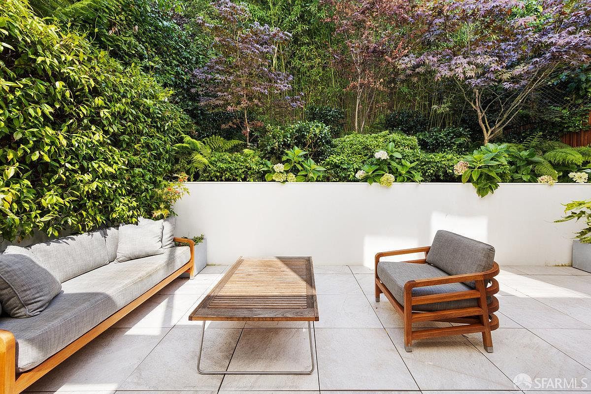 This serene outdoor patio features a modern aesthetic with light-toned stone pavers and a clean, white retaining wall. The space is furnished with a wooden sofa and matching armchair, both topped with neutral gray cushions, arranged around a low-profile wooden coffee table. Lush greenery and flowering shrubs provide a vibrant, private backdrop, creating a peaceful and inviting atmosphere for relaxation.