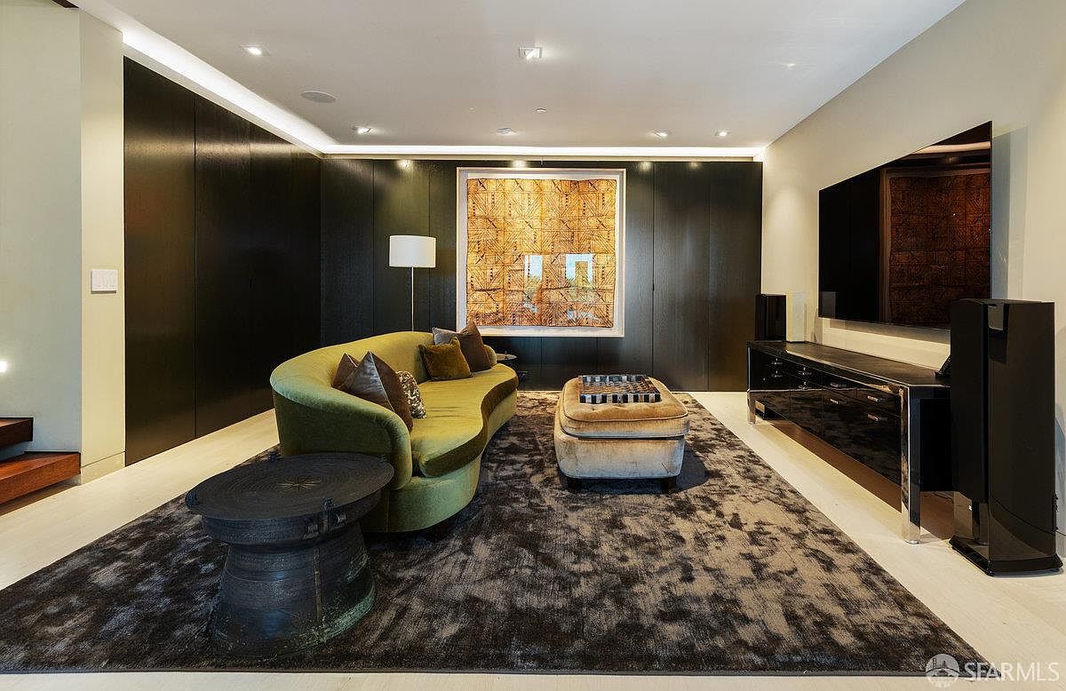 This sophisticated living room features a curved olive-green sofa centered on a large, textured area rug, complemented by a velvet ottoman and a dark circular side table. The room is anchored by a striking, large-scale geometric art piece mounted on a dark, paneled wall, while a sleek media console sits beneath a wall-mounted television. The space is illuminated by recessed lighting and a minimalist floor lamp, creating a moody, contemporary atmosphere.