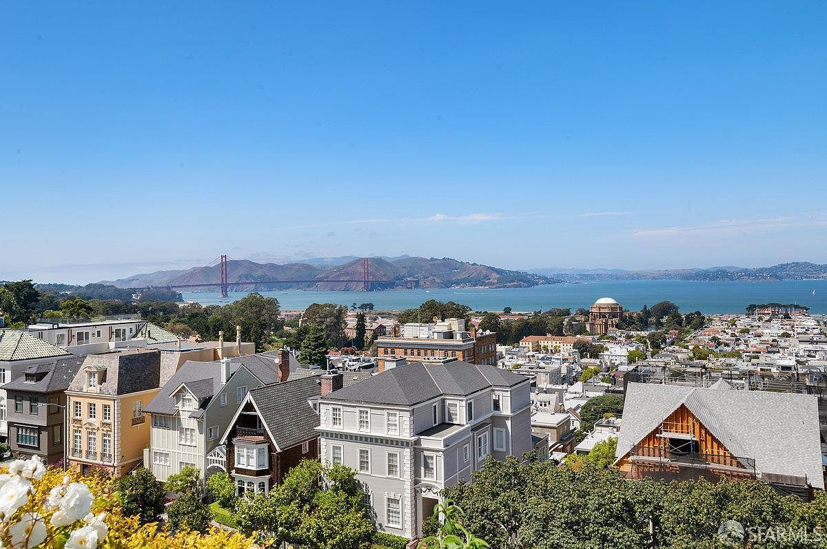 This elevated, wide-angle shot captures a stunning panoramic view of a residential neighborhood in San Francisco, featuring the iconic Golden Gate Bridge in the distance. The foreground showcases a collection of classic, multi-story homes with varied architectural styles, including gabled roofs and traditional siding, nestled among lush green trees. The clear blue sky and expansive bay water create a serene and highly desirable setting, emphasizing the property's prime location and scenic outlook.