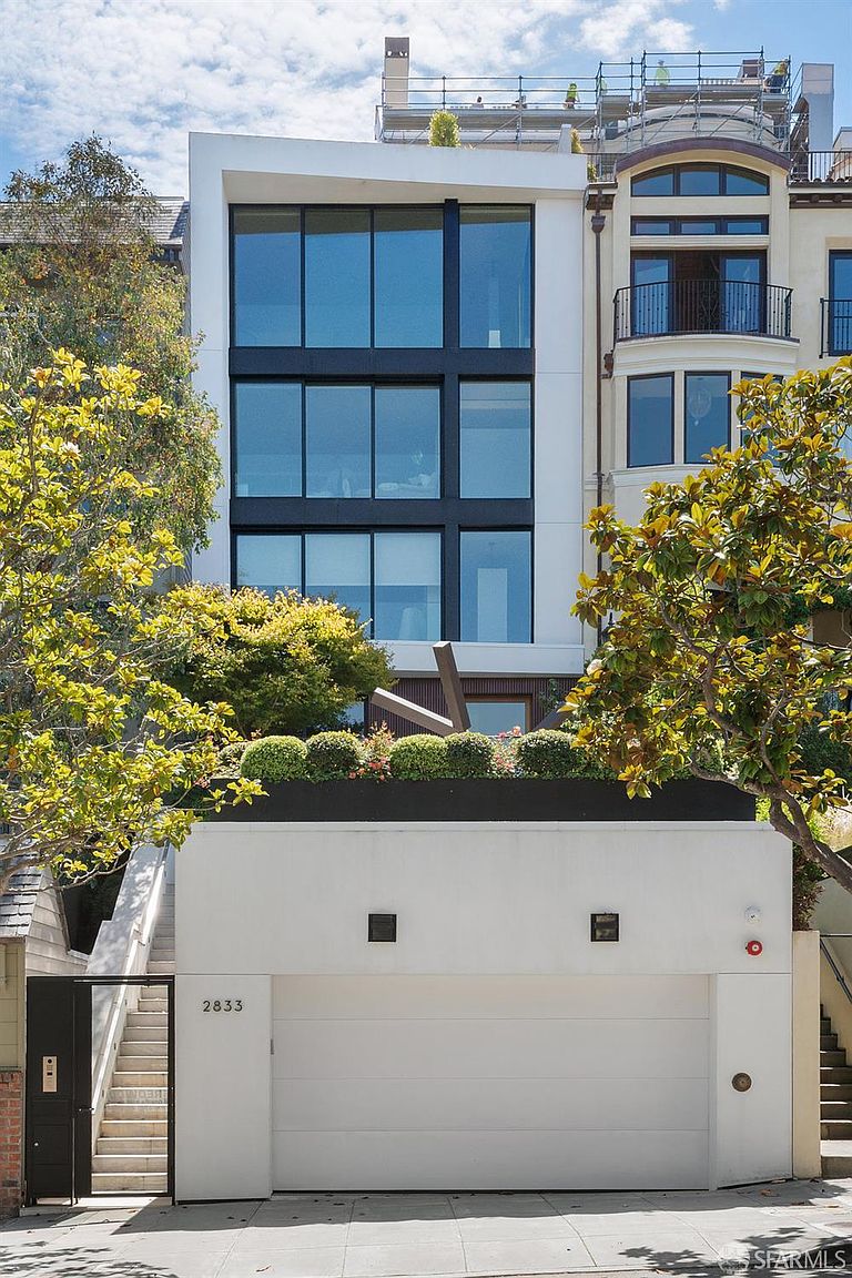This modern, multi-story residence features a striking facade with large, grid-patterned windows and a clean, white exterior finish. A private garage occupies the ground level, topped by a landscaped terrace with manicured shrubs and a sculptural art piece. The property is framed by mature trees and adjacent to a more traditional building, creating a distinct contrast between contemporary and classic architectural styles.