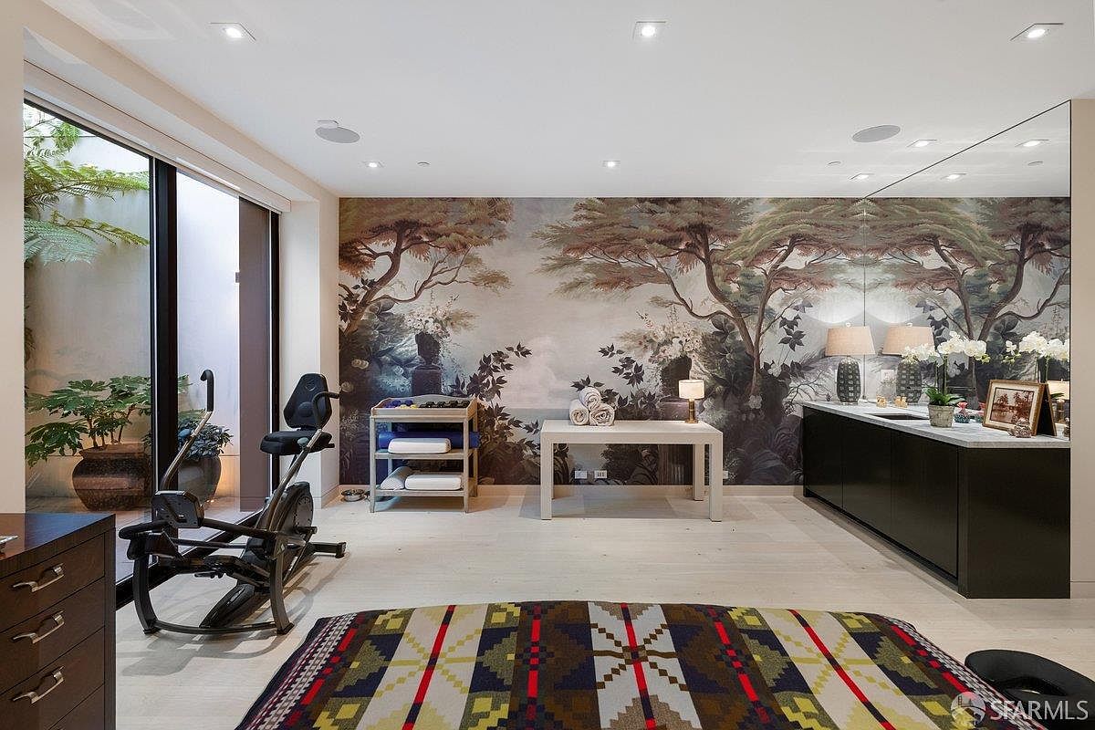 This home gym features a striking mural wall depicting a lush, classical landscape, creating a serene atmosphere for exercise. The room is equipped with a stationary bike, a minimalist console table, and a storage rack, all set against light-toned wood flooring. Large glass doors on the left provide a view into a private, landscaped courtyard, while a large mirror on the right wall enhances the sense of space.
