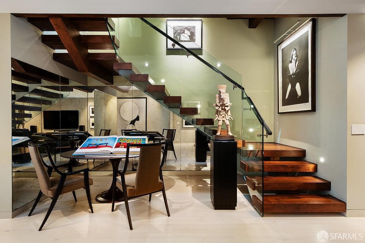 This sophisticated interior space features a striking floating staircase with dark wood treads and a glass railing, creating an open and airy aesthetic. A circular dining table sits beneath the stairs, complemented by a mirrored wall that enhances the sense of depth and light. The area is decorated with curated artwork and a sculptural piece on a pedestal, reflecting a modern, gallery-like design style.