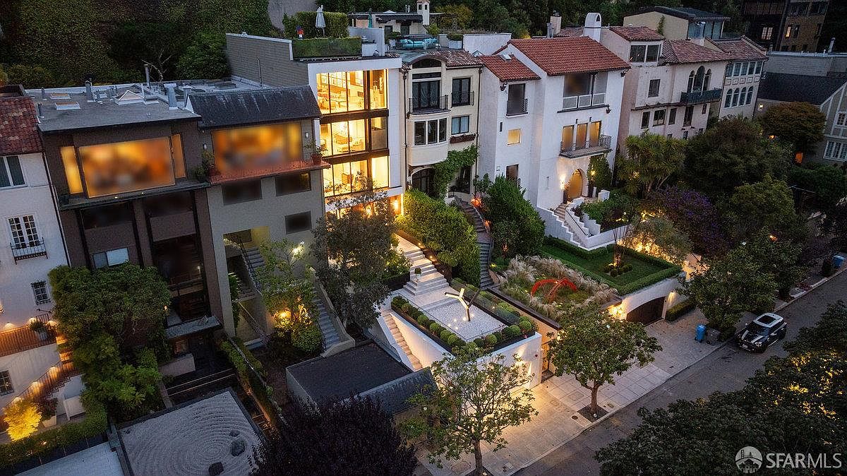 This high-angle aerial view captures a row of sophisticated, multi-story urban residences at dusk, highlighting their unique architectural designs and tiered landscaping. The central property features a striking glass-fronted facade that glows warmly, contrasting with the surrounding homes and the meticulously manicured terraced gardens below. The perspective emphasizes the seamless integration of modern luxury living with outdoor spaces, including stone pathways and sculptural garden elements, set against a quiet neighborhood street.