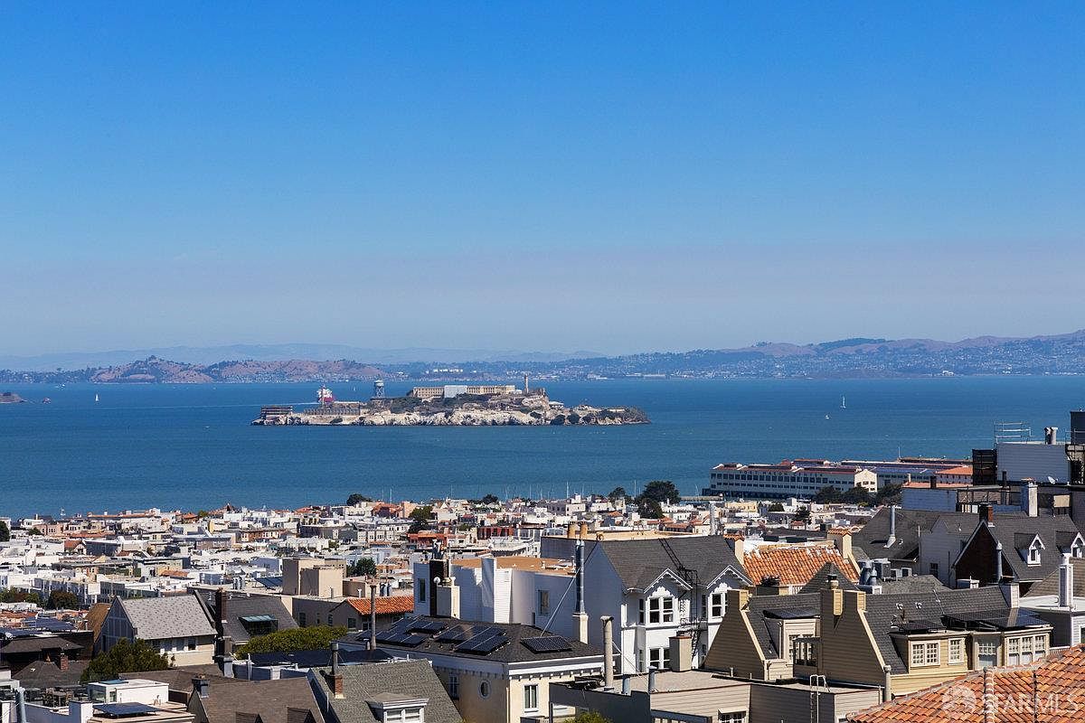 This elevated perspective captures a sweeping panoramic view of a dense urban neighborhood leading down to the San Francisco Bay. The iconic Alcatraz Island is prominently centered in the distance, framed by a clear blue sky and rolling hills on the horizon. The foreground showcases the intricate rooftops and architectural character of the city's residential landscape.