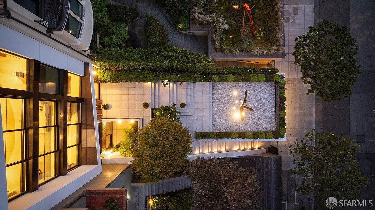 This high-angle aerial view captures a sophisticated urban outdoor space, featuring a multi-level terrace with stone paving, manicured hedges, and a central sculpture. The composition highlights a modern building with large windows on the left, contrasting with the geometric landscaping and ambient evening lighting below. The perspective provides a comprehensive look at the property's architectural layout and private outdoor living areas.