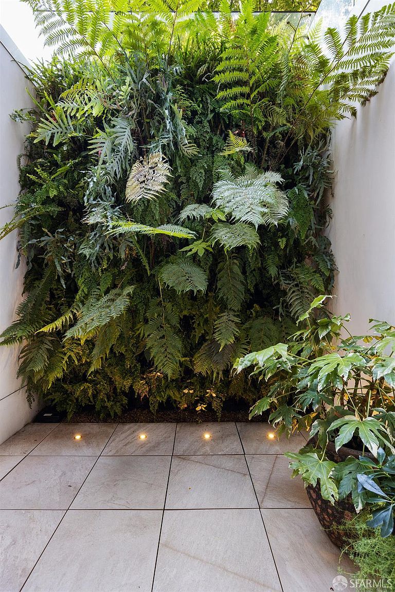 This serene outdoor space features a striking vertical garden wall densely planted with various ferns, creating a lush, living backdrop. The ground is paved with large, light-toned square tiles, accented by recessed ground-level lighting that casts a warm glow upward. A potted plant with broad, palmate leaves sits in the foreground, adding depth to this tranquil, modern courtyard setting.
