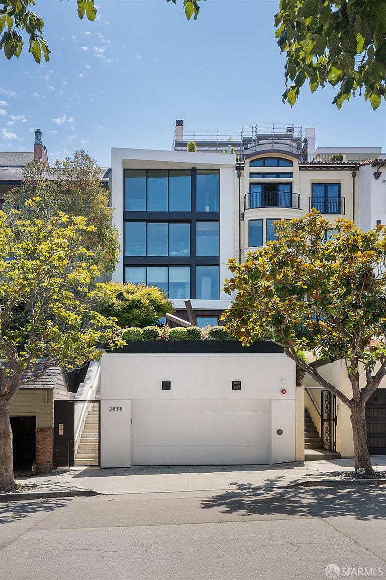 This front-facing view captures a modern, multi-story residence characterized by a striking contrast between a contemporary glass-fronted structure and a more traditional neighboring building. The property features a prominent white garage door at street level, topped by a landscaped terrace with lush greenery. The upper levels showcase large, grid-patterned windows that reflect the sky, creating a sophisticated and architectural aesthetic.