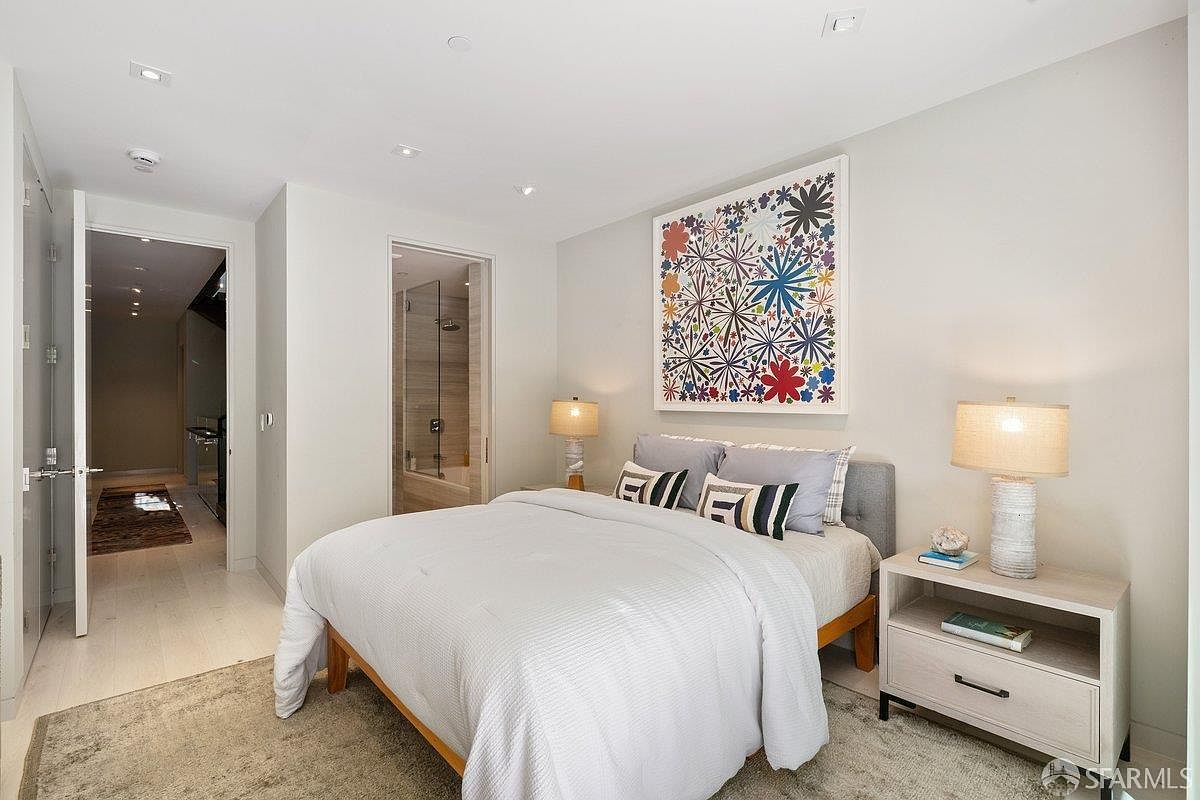 This modern primary bedroom features a neutral color palette with light wood flooring and a textured area rug. A central bed is adorned with patterned accent pillows and sits beneath a vibrant, abstract floral art piece, flanked by two matching bedside tables with textured lamps. The room includes a doorway leading to an en-suite bathroom, creating a clean and cohesive living space.