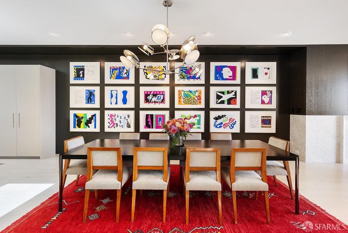 This sophisticated dining room features a long, dark-toned table surrounded by eight light-upholstered chairs with wood frames, set atop a vibrant red patterned rug. The focal point is a dark accent wall adorned with a gallery display of eighteen colorful, framed abstract prints arranged in a grid. A modern, multi-armed chandelier hangs above the table, which is centered with a large, fresh floral arrangement, creating an elegant and artistic atmosphere.