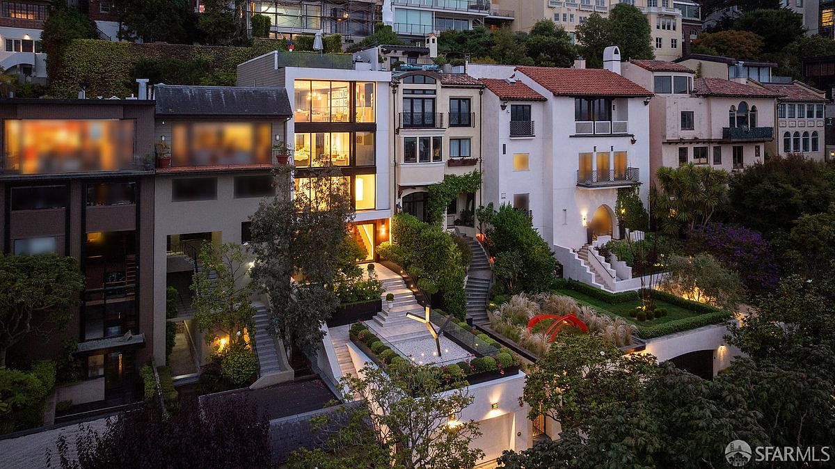 This elevated perspective captures a row of multi-story hillside residences in San Francisco at dusk, showcasing a blend of modern and Mediterranean architectural styles. The central home features a striking glass-fronted facade with illuminated interiors, tiered stone staircases, and meticulously landscaped garden terraces. The scene is framed by lush greenery and warm exterior lighting, creating a sophisticated and inviting atmosphere.