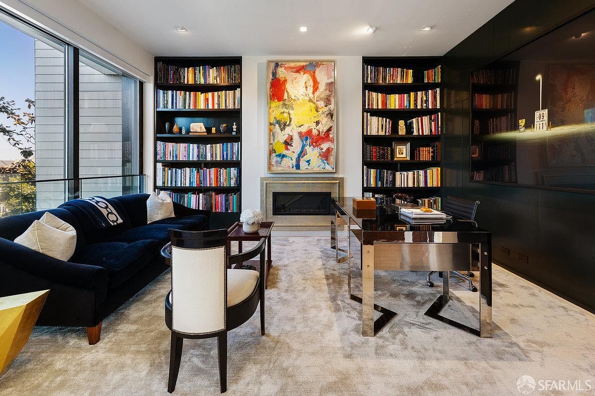 This sophisticated home office features a sleek, modern desk with a metallic base, paired with a dark-toned sofa and a contemporary accent chair. The room is anchored by two tall, built-in bookshelves flanking a central fireplace with a vibrant abstract painting above it. Large windows to the left provide ample natural light and a view of the outdoors, while the overall aesthetic is refined and professional.