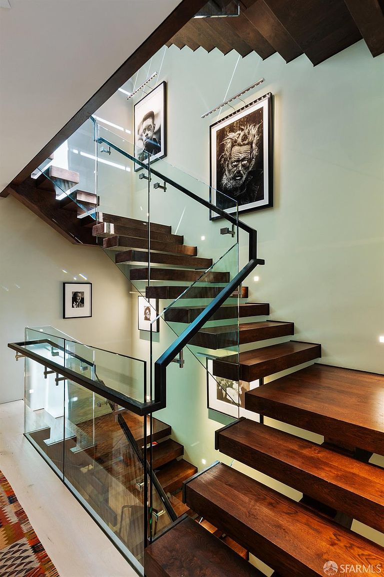 This striking staircase features thick, dark wood treads that appear to float, complemented by a modern glass railing and a sleek black metal handrail. The light-colored walls are adorned with framed black-and-white portraits, creating a gallery-like atmosphere. The perspective is captured from a high angle, emphasizing the architectural geometry and the open, airy design of the stairwell.
