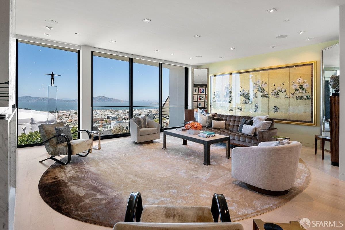 This sophisticated living room features a large, circular area rug anchoring a seating arrangement of a plush sofa and several armchairs. The space is defined by expansive, wall-to-wall windows that provide a panoramic view of the city and bay, while a large, ornate decorative screen serves as a focal point on the adjacent wall. The room is bathed in natural light, creating an airy and luxurious atmosphere suitable for high-end urban living.