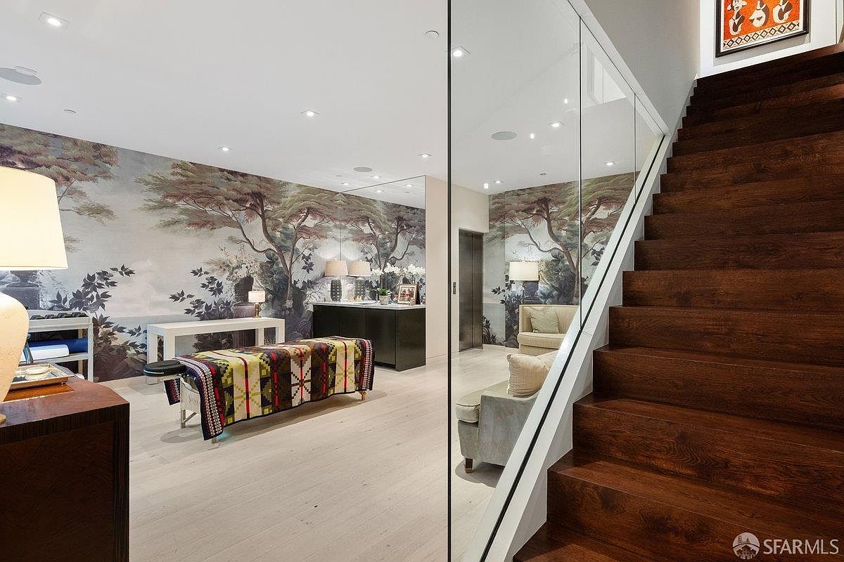 This interior view captures a sophisticated transition space featuring a striking wooden staircase with dark, rich treads. To the left, a mirrored wall reflects a seating area adorned with mural-style wallpaper depicting a serene landscape, creating an illusion of expanded space. The area is illuminated by recessed lighting and a stylish lamp, contributing to a refined and artistic atmosphere.