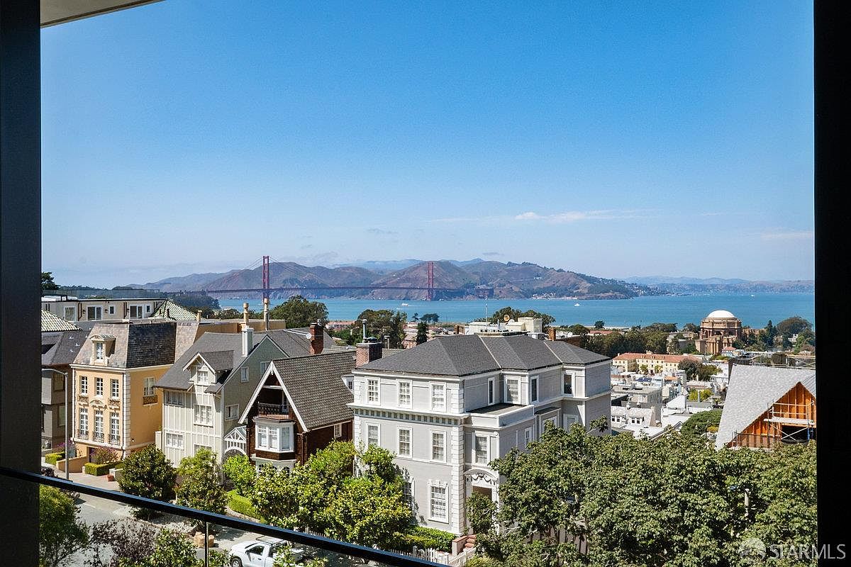 This image is captured from an elevated balcony, offering a sweeping, cinematic perspective of a residential neighborhood in San Francisco. The view prominently features the iconic Golden Gate Bridge spanning the bay in the distance, framed by lush trees and classic architectural homes in the foreground. The clear blue sky and bright daylight emphasize the prime location and the expansive, picturesque scenery.