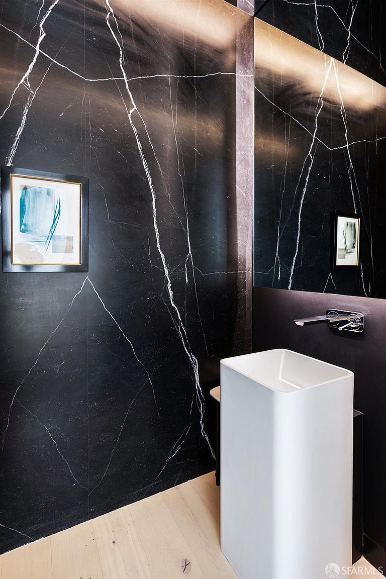 This modern powder room features a striking wall clad in dark, white-veined stone that creates a dramatic backdrop. A minimalist, white pedestal sink is paired with a sleek, wall-mounted chrome faucet, while a framed piece of abstract art adds a touch of color to the space. The room is finished with light-toned wood flooring, providing a sharp contrast to the dark wall and creating a sophisticated, high-end aesthetic.