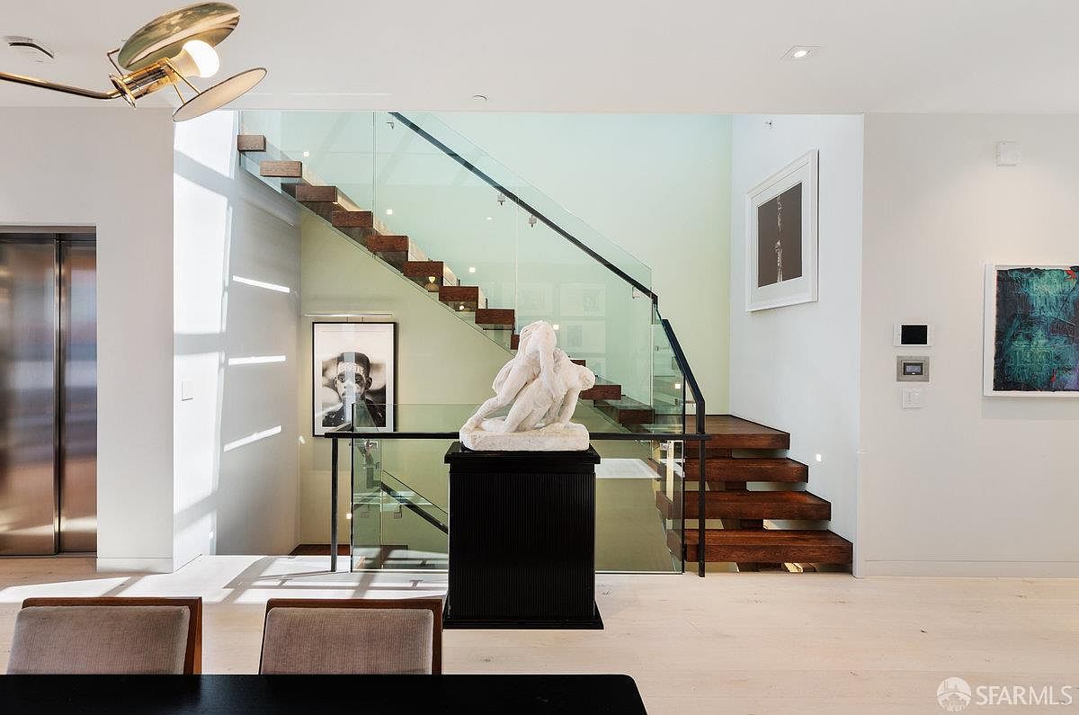 This sophisticated interior space features a dramatic floating staircase with dark wood treads and a glass railing, serving as a central architectural focal point. A white sculpture sits atop a dark pedestal in the foreground, positioned in front of the stairs, while modern artwork adorns the light-colored walls. The perspective is eye-level, capturing the clean lines and contemporary aesthetic of the home's open-plan design.