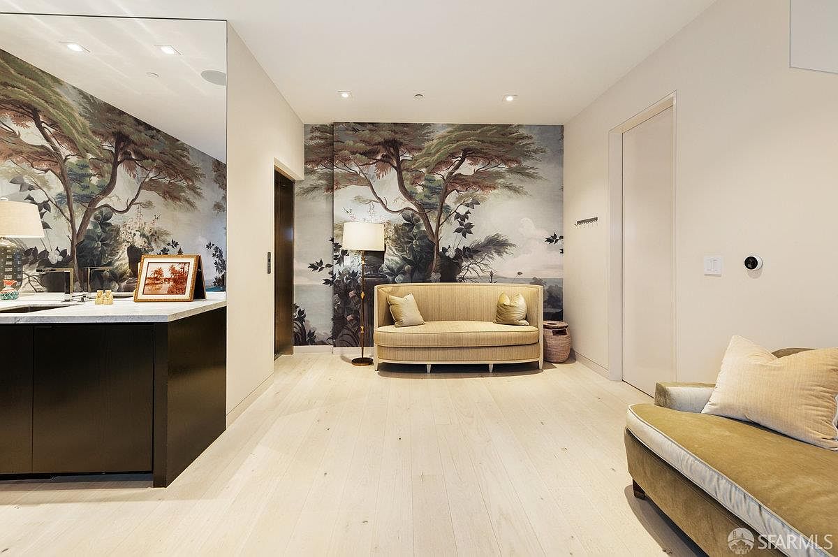 This sophisticated interior space features a light-toned wood floor and a striking mural wall depicting a serene landscape with trees. A neutral-toned sofa sits centered against the mural, complemented by a floor lamp and a matching sofa in the foreground. The room includes a large mirror on the left wall, reflecting the space and adding depth, while recessed lighting provides a clean, modern ambiance.