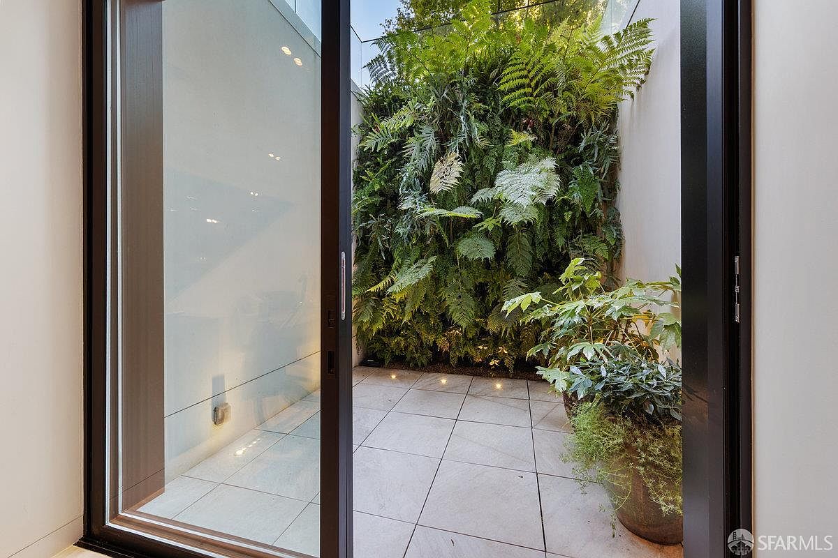 This image captures a modern, private courtyard or light well viewed through a large sliding glass door. The space features light-colored square floor tiles and a lush, vertical living wall filled with various ferns and greenery. The perspective from inside looking out emphasizes the seamless transition between the interior living space and the serene, nature-focused outdoor retreat.