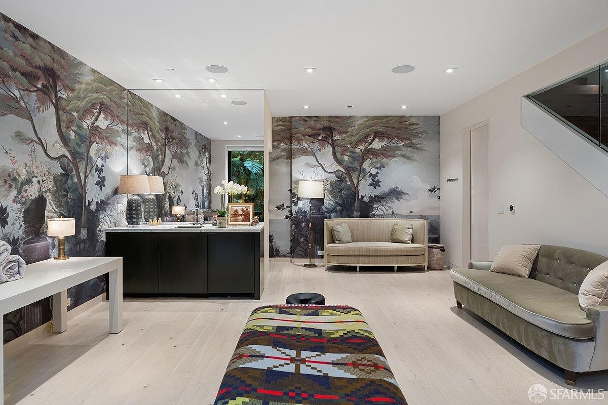 This sophisticated interior space features a mural-style wallpaper depicting a lush forest scene, creating a serene and artistic atmosphere. The room is furnished with two distinct seating areas, including a curved sofa and a patterned bench, set against light-toned wood flooring. A sleek dark-toned wet bar with a mirror backsplash adds a functional and modern touch to this elegant lounge area.