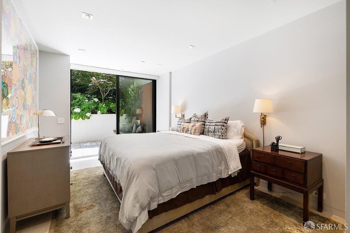 This primary bedroom features a neutral color palette with a large bed centered against a plain wall, flanked by two wall-mounted lamps. To the left, a dresser sits beneath a vibrant, multi-colored circular art piece, while a large sliding glass door provides access to a private outdoor patio area. The room is carpeted and maintains a clean, contemporary aesthetic with ample natural light.