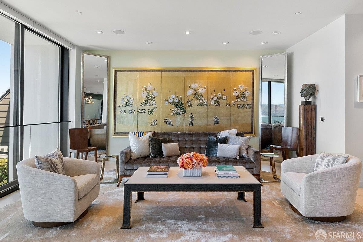 This sophisticated living room features a plush, tufted velvet sofa centered beneath a large, ornate gold-framed screen depicting floral motifs. The space is anchored by a light-toned coffee table and two curved, upholstered armchairs, all resting on a textured area rug. Large windows on the left and right provide natural light and views, while tall mirrors flanking the central artwork enhance the sense of depth and elegance in this refined seating area.
