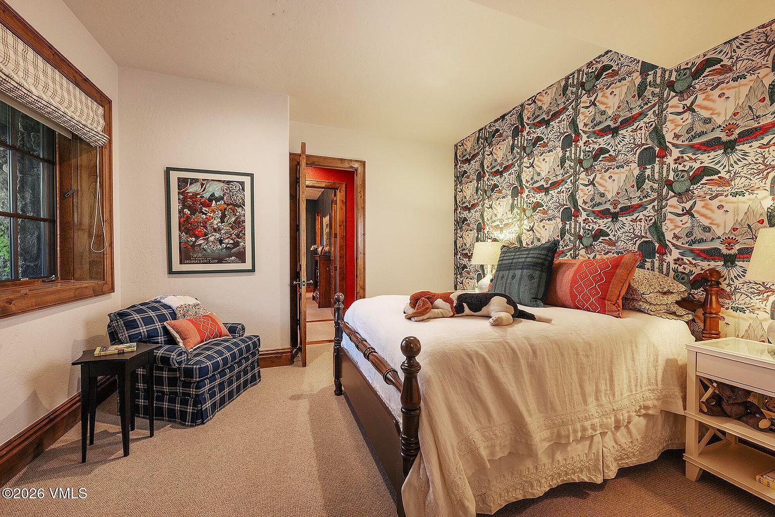 This cozy bedroom features a distinctive patterned wallpaper with an avian motif behind a bed dressed in neutral linens and accented with colorful throw pillows. A plaid armchair sits in the corner near a window with wood trim, while a framed piece of art hangs on the adjacent wall. The room includes a wooden bed frame and a small side table, with a doorway leading into a hallway with red-painted walls.