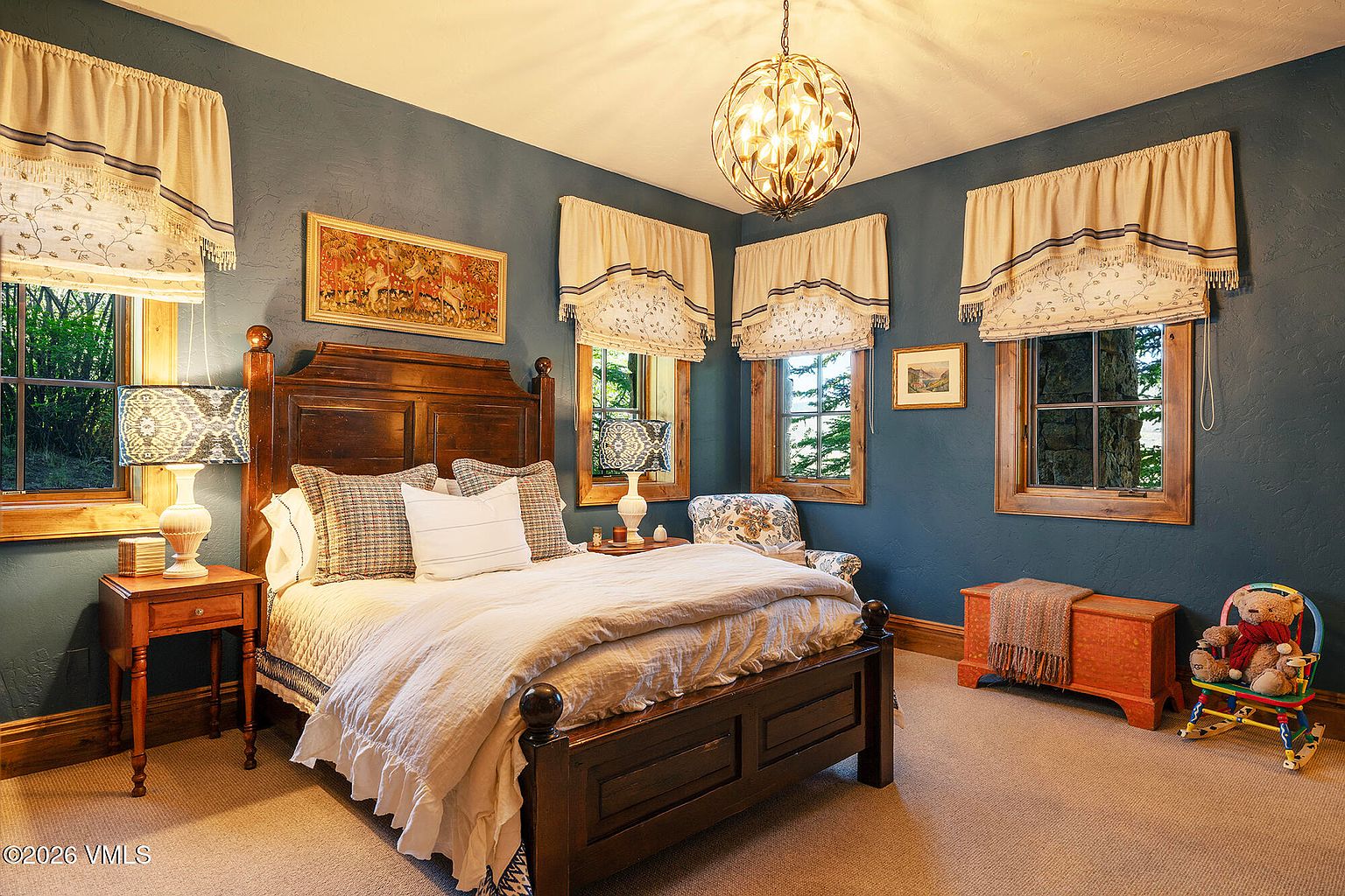 This cozy bedroom features deep blue walls, a dark wood bed frame, and a matching nightstand with a patterned lamp. The room is accented with patterned window valances, a decorative chandelier, and a small wooden chest at the foot of the bed. A child's rocking chair with a teddy bear adds a charming touch to this well-appointed space.