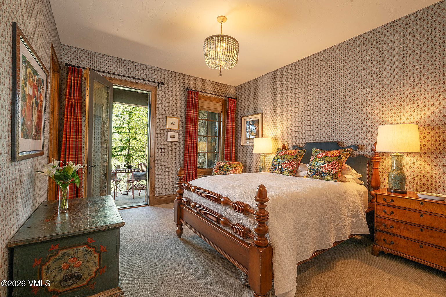 This cozy bedroom features a traditional aesthetic with patterned wallpaper, a wooden bed frame, and a matching wooden dresser. A glass door opens to a private balcony, allowing natural light to fill the room, while a decorative chandelier provides warm overhead lighting. The space is accented by floral-patterned throw pillows and two bedside lamps, creating a comfortable and inviting atmosphere.